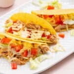 Three chicken tacos with shredded cheese, lettuce, and diced tomatoes on a white plate.
