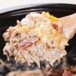 A wooden spoon scooping out cheesy chicken and bacon from a slow cooker.