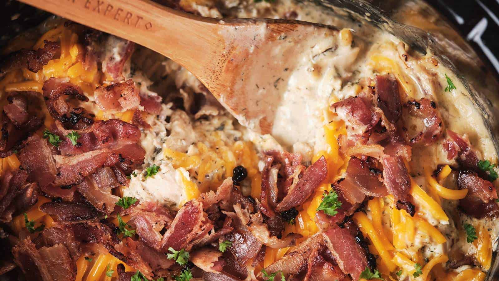 Creamy, cheesy chicken dish garnished with crispy bacon pieces and parsley, stirred with a wooden spoon.