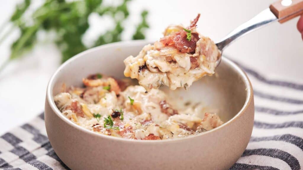 A fork lifts a serving of creamy chicken topped with bacon and garnished with herbs from a ceramic bowl.