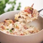 A fork lifts a serving of creamy chicken topped with bacon and garnished with herbs from a ceramic bowl.
