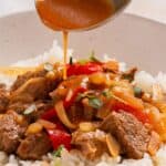 Sauce being poured over a bowl of beef stir fry with bell peppers and onions on a bed of white rice.