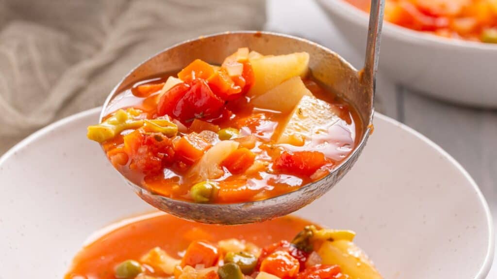 Ladle serving vegetable soup with tomatoes, potatoes, and green beans into a white bowl.