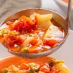 Ladle serving vegetable soup with tomatoes, potatoes, and green beans into a white bowl.