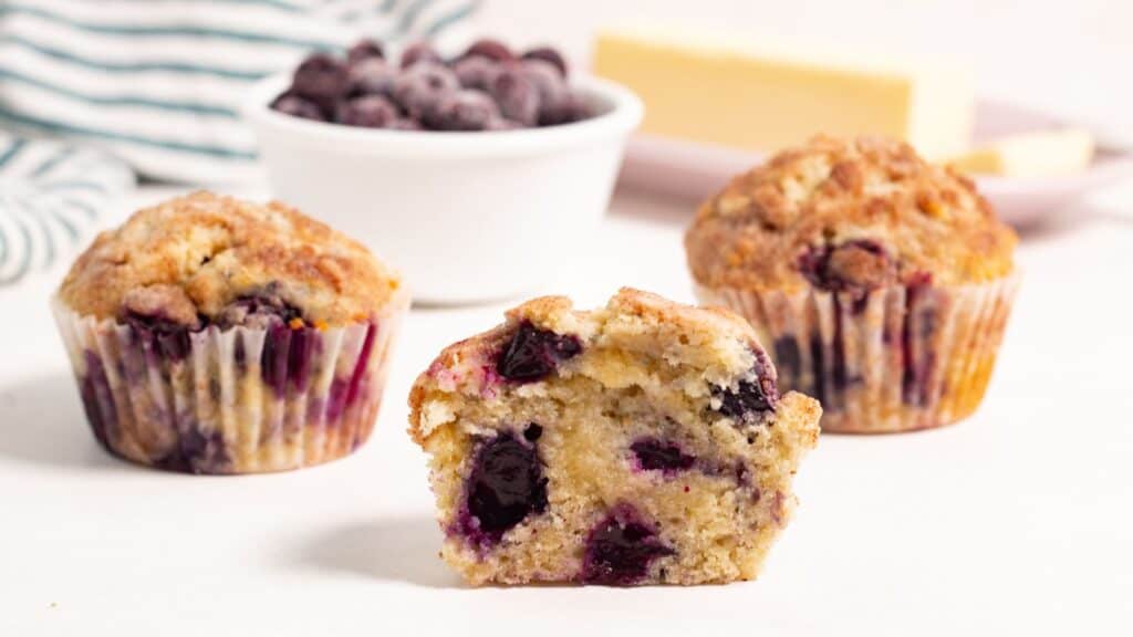 Two blueberry muffins, one halved, with fresh blueberries and a slice of butter in the background.