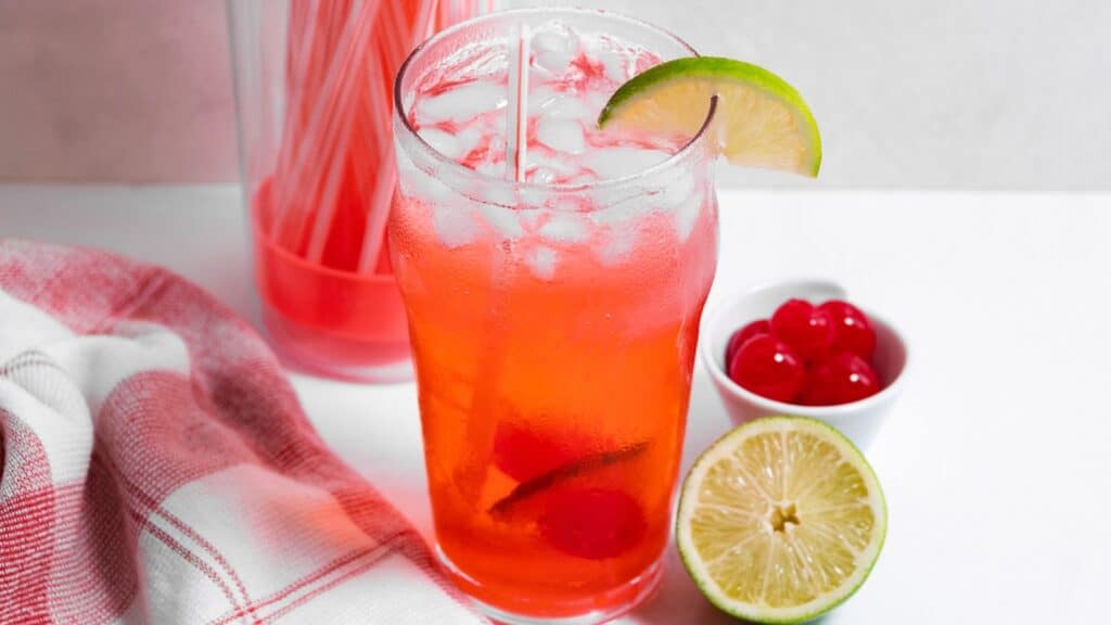 A glass of cherry limeade with ice, garnished with a lime slice, served next to a bowl of cherries and a pitcher of more limeade.