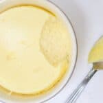 A serving of fresh, creamy cheesecake with a spoon taking a scoop out, placed on a white surface.