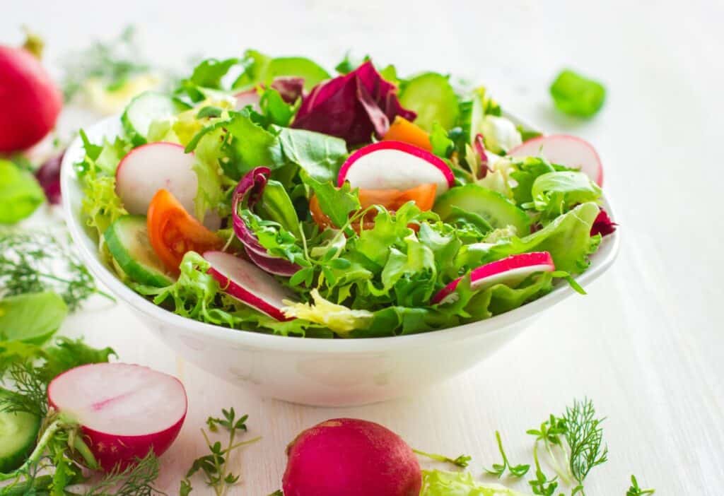 A fresh mixed vegetable salad with lettuce, radishes, cucumber, and tomato in a white bowl.