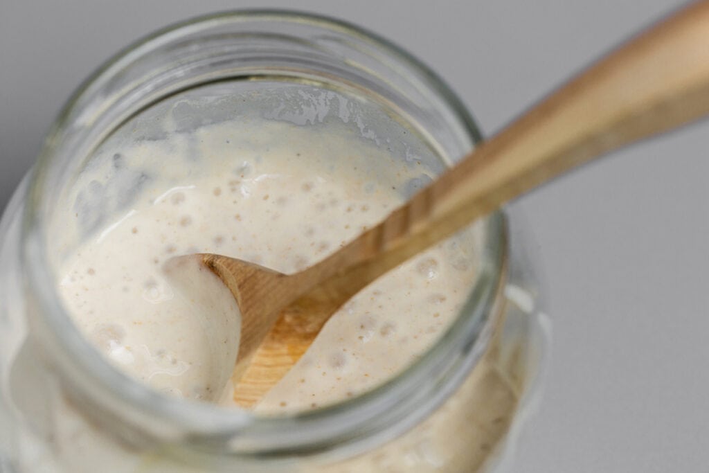 A close-up view of feeding sourdough starter in a glass jar with a wooden stirrer.