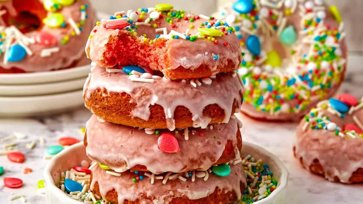 A stack of colorful, glazed doughnuts with sprinkles, displayed on a white surface surrounded by scattered candies.