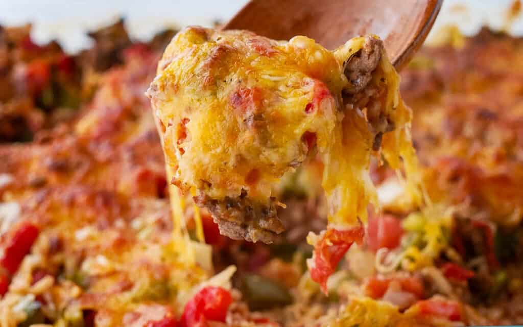 A spoon lifting a cheesy portion of baked casserole with visible layers of meat and vegetables.