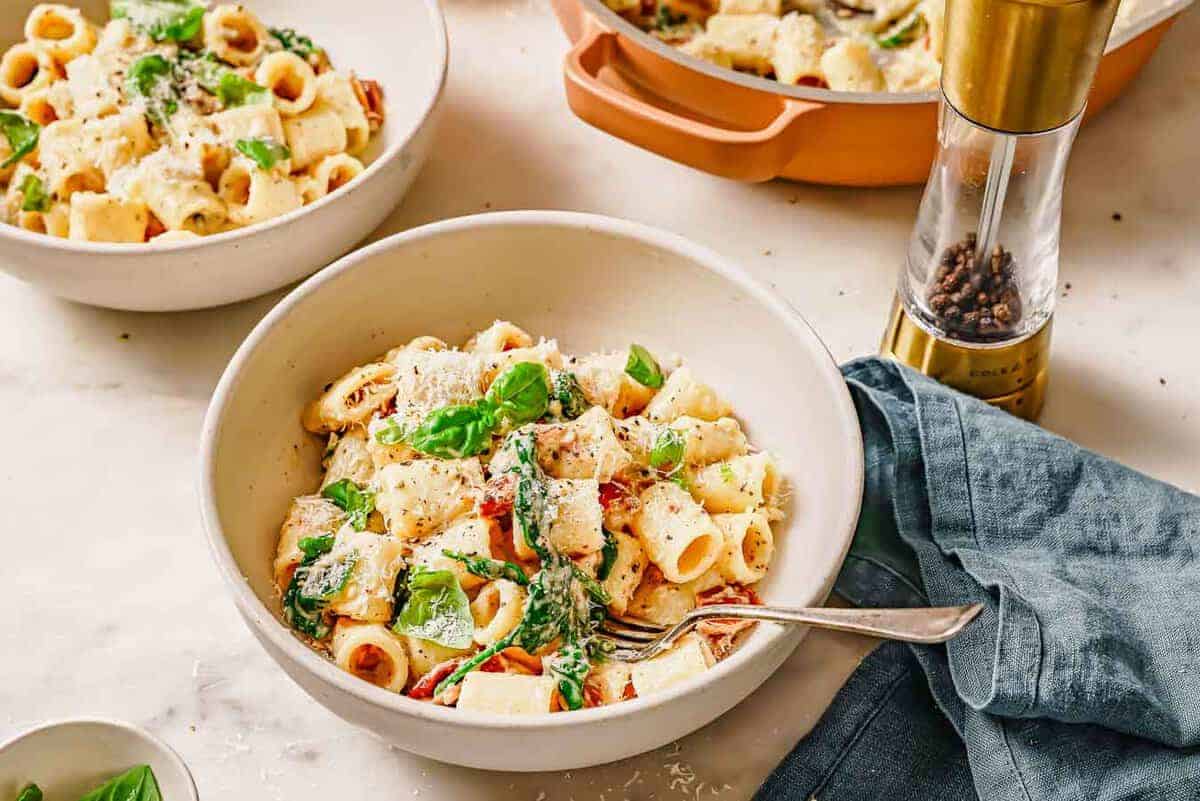 A bowl of pasta with vegetables and cheese garnished with basil on a table next to a pepper mill and a linen napkin.