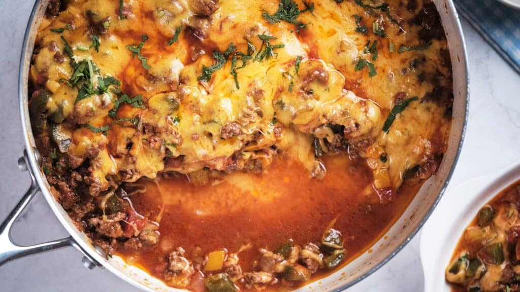 A skillet with a cheesy ground beef and vegetable casserole, sprinkled with herbs, displayed on a table.