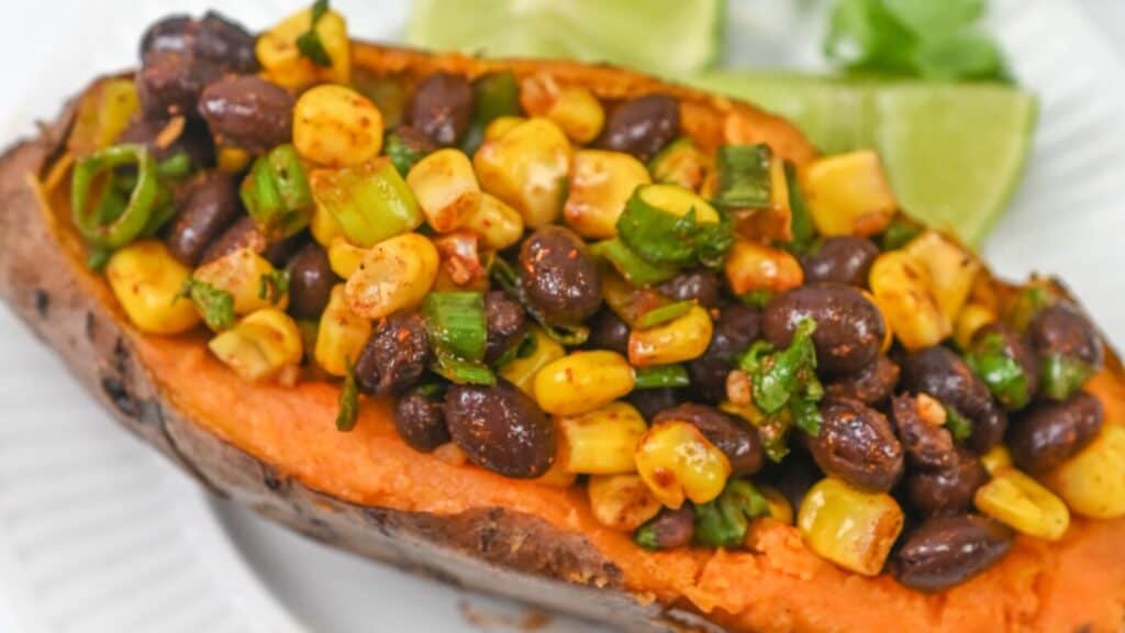 A baked sweet potato topped with sautéed black beans, corn, and green onions, garnished with lime wedges.