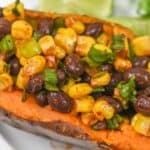 A baked sweet potato topped with sautéed black beans, corn, and green onions, garnished with lime wedges.