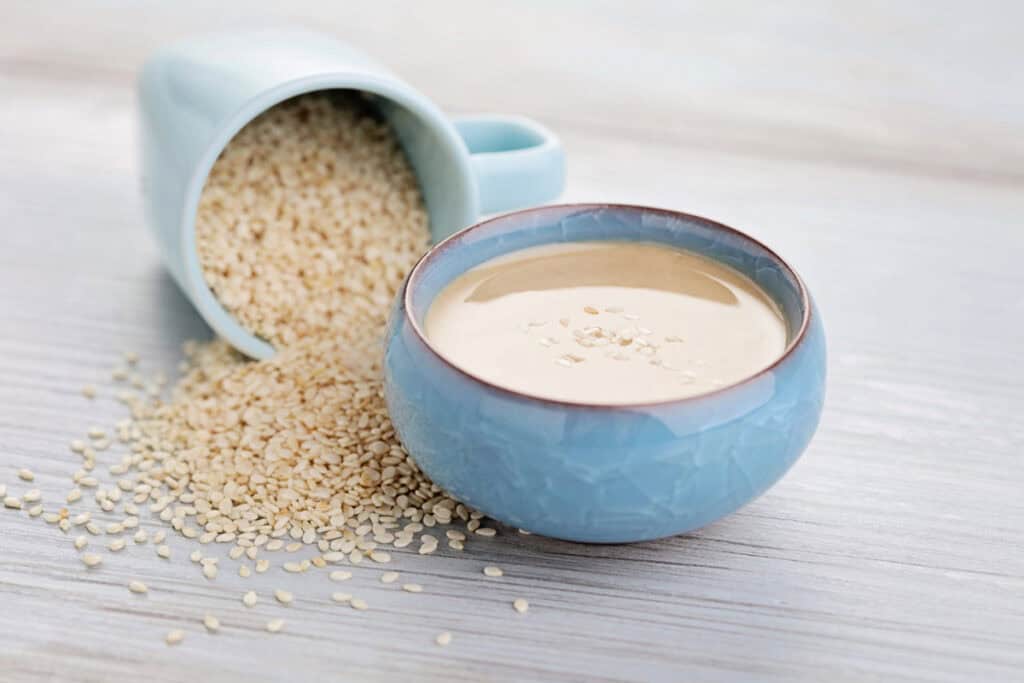 A bowl of creamy tahini made from sesame seeds, with a spilled cup of sesame seeds beside it on a wooden surface.