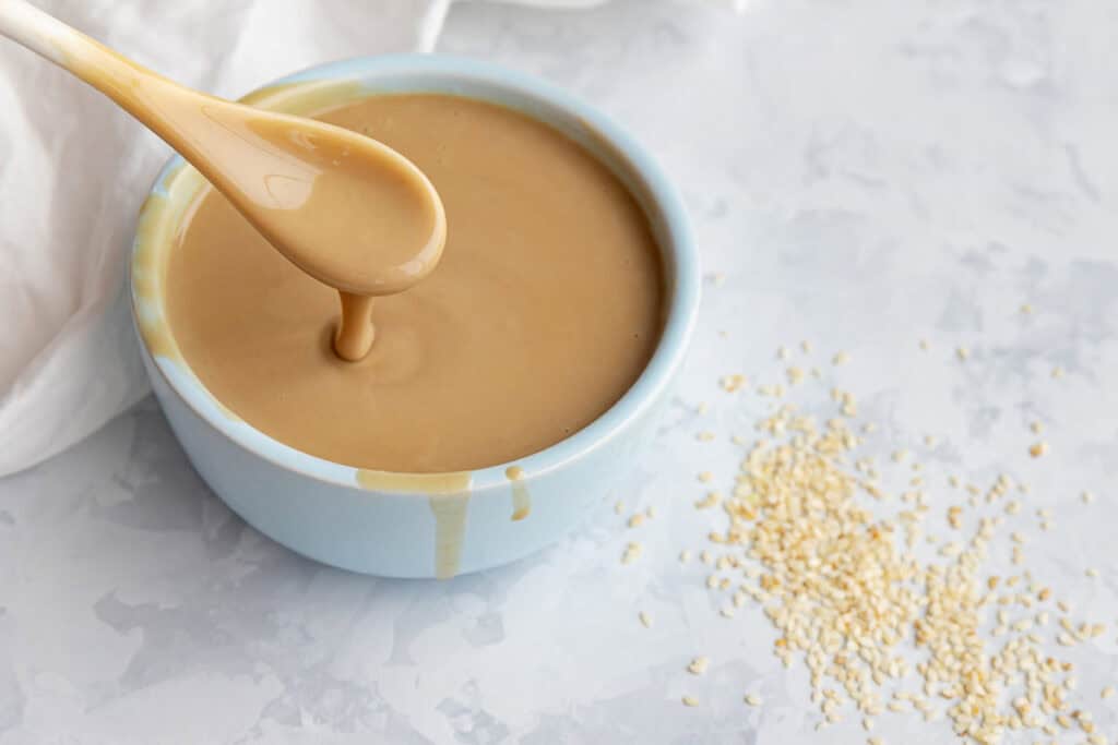 A light blue bowl filled with creamy tahini on a marble surface, a spoon dripping tahini above it, and sesame seeds scattered nearby.