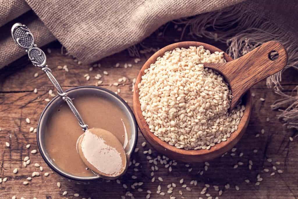 A wooden bowl of sesame seeds with a scoop, next to a bowl of tahini and a spoon, on a rustic wooden table.