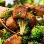 Roasted broccoli and tofu cubes in a wooden spoon, garnished with sesame seeds and green onions.