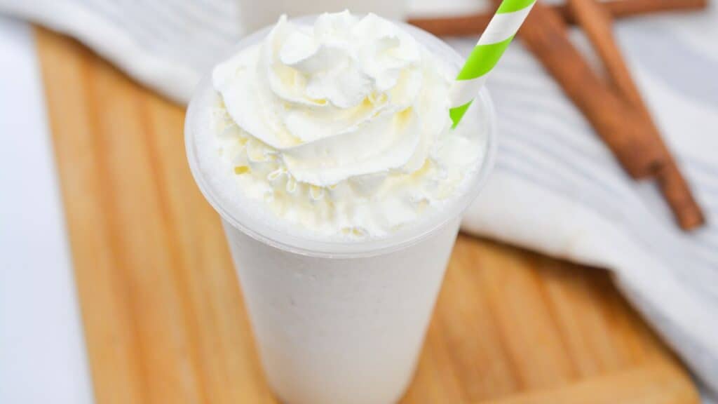 A whipped cream-topped milkshake with a striped straw on a wooden surface with cinnamon sticks beside it.