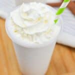 A whipped cream-topped milkshake with a striped straw on a wooden surface with cinnamon sticks beside it.