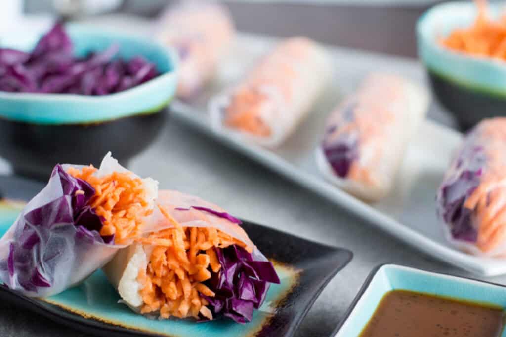 A variety of fresh spring rolls with carrots and purple cabbage, served with a dipping sauce on a modern ceramic plate.