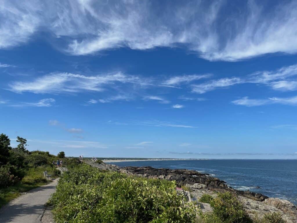 Coastal path in Maine with lush greenery on the left and rocky shore on the right under a blue sky with sweeping clouds.