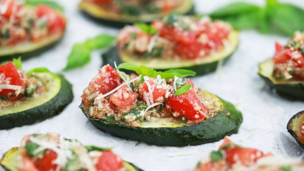 Baked zucchini bruschetta topped with chopped tomatoes, herbs, and grated cheese on a parchment-lined tray.