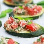 Baked zucchini bruschetta topped with chopped tomatoes, herbs, and grated cheese on a parchment-lined tray.