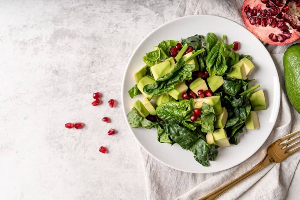 A white plate with a fresh avocado, spinach, and pomegranate salad, with a gold fork and a pomegranate on the side, on a light gray surface.