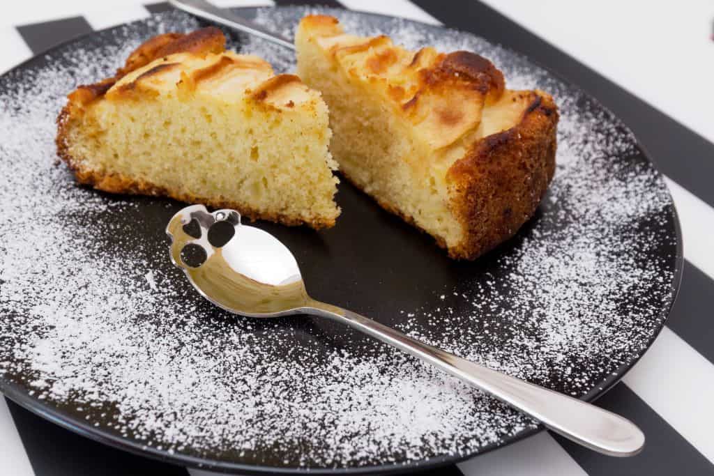 A black plate with two slices of sponge cake dusted with powdered sugar and a silver spoon with a decorative design on the handle, on a striped black and white surface.