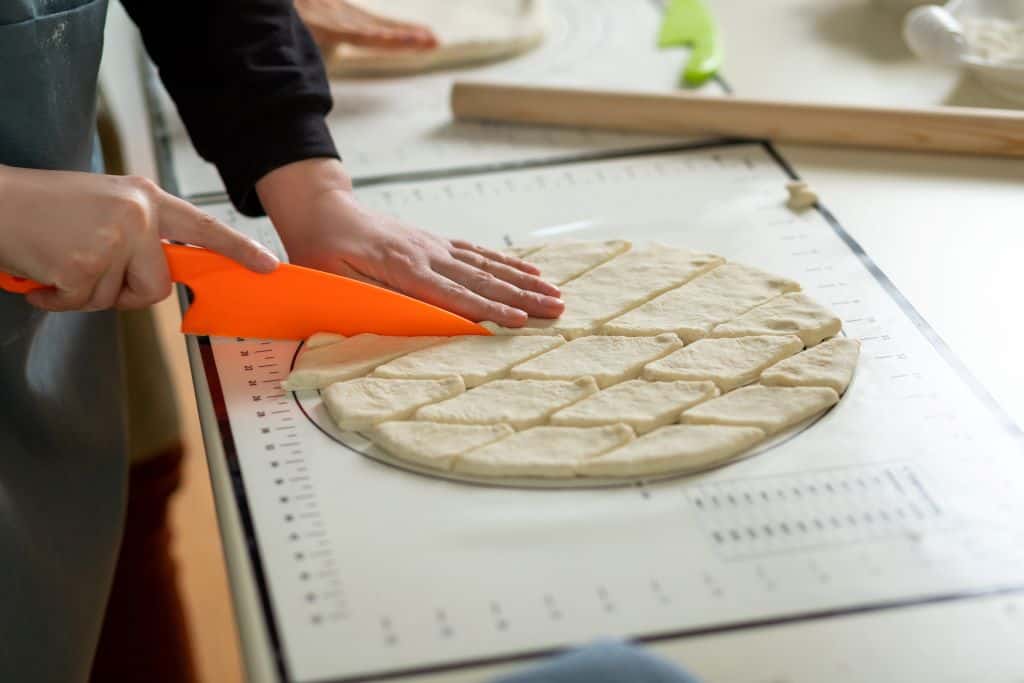 Close-up of hands using an orange knife to slice dough into diamond shapes on a pastry board.