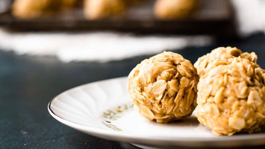 Two oatmeal peanut butter balls sit on a white plate, with more in the background on a dark surface.