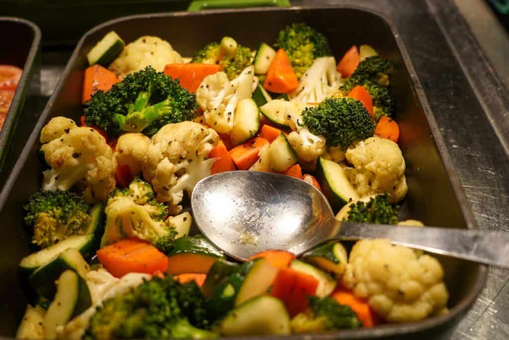 A tray of steamed vegetables including broccoli, cauliflower, and carrots, with a serving spoon, displayed under warm lighting.