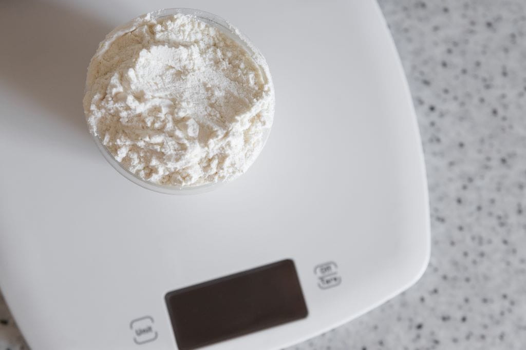 A clear plastic cup filled with white flour is sitting on a digital kitchen scale on a speckled countertop.