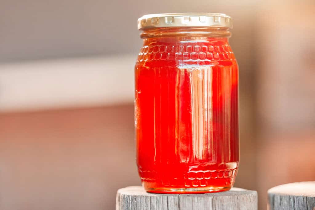 A clear glass jar filled with red syrup, sealed with a gold lid, standing on a wooden surface.