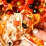 A hand holds a chip topped with a layer of sour cream, tomatoes, black olives, green onions, and shredded cheese above a platter of loaded nachos.