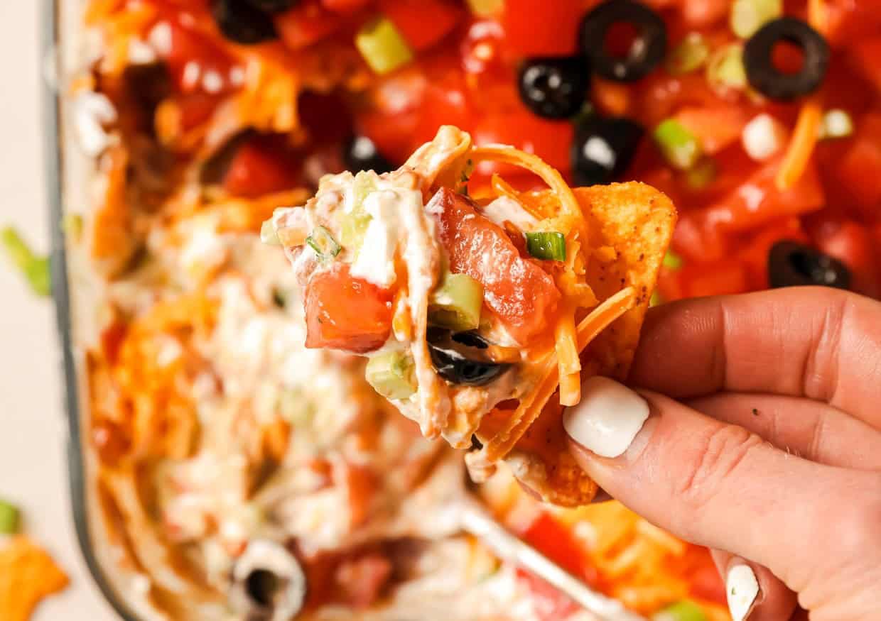 A hand holds a chip topped with a layer of sour cream, tomatoes, black olives, green onions, and shredded cheese above a platter of loaded nachos.