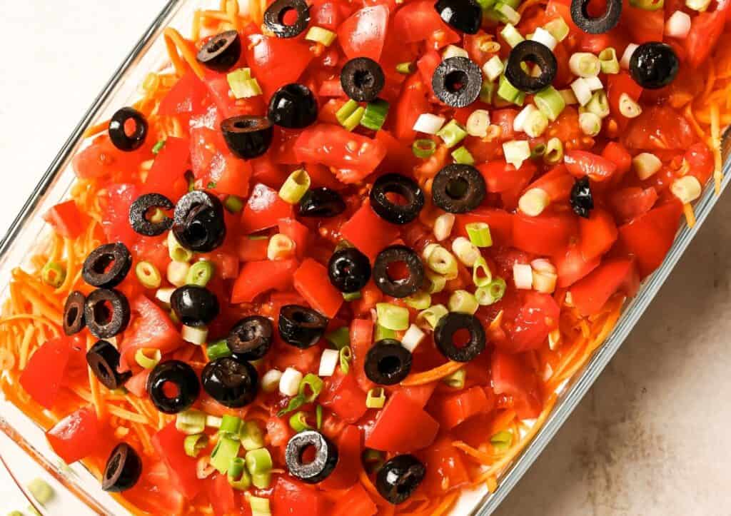 A glass dish filled with a layered salad featuring shredded cheese, diced tomatoes, sliced black olives, and chopped green onions.