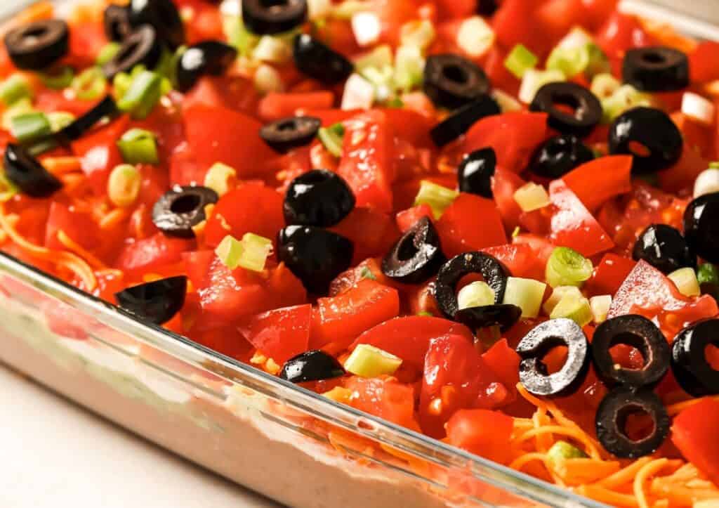 A close-up of a layered taco dip topped with chopped tomatoes, black olives, green onions, and shredded cheese in a rectangular glass dish.