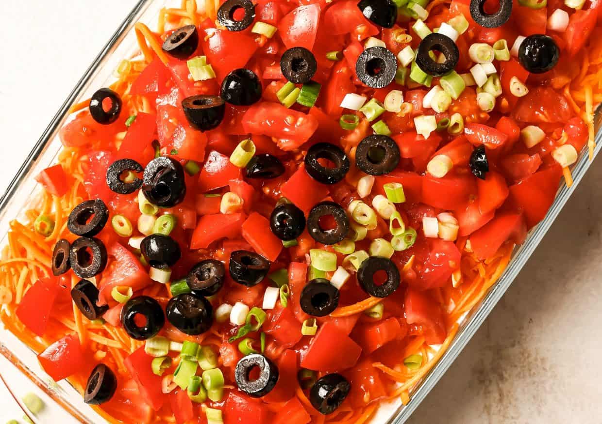 A baking dish with a layer of sour cream, tomatoes, black olives, green onions, and shredded cheese above a platter of loaded nachos.