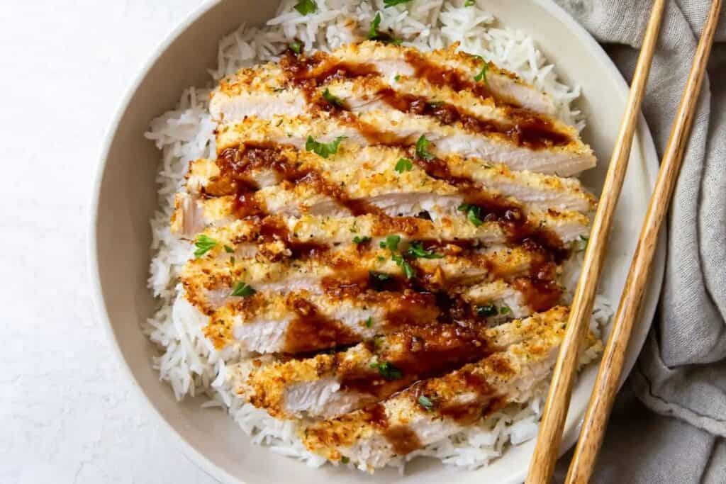 Slices of breaded and seasoned chicken breast placed on a bed of white rice, drizzled with sauce, and garnished with herbs. Chopsticks are resting on the side of the bowl.