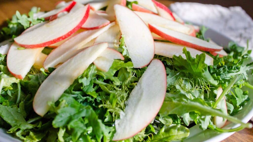 A salad consisting of mixed greens topped with thinly sliced red apple pieces on a white plate.
