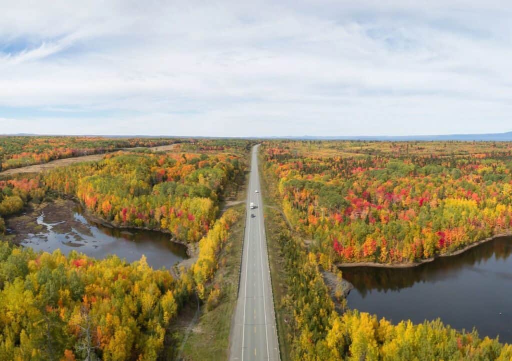 A straight road runs through a vibrant autumn forest with trees in various shades of green, yellow, orange, and red. Two lakes are visible on either side of the road under a partly cloudy sky.