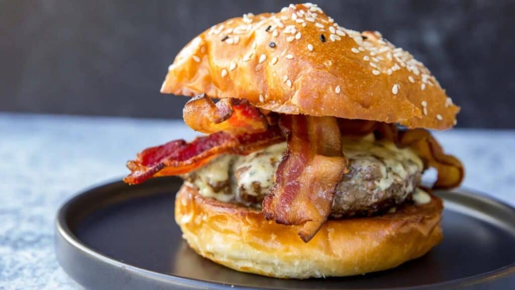 A bacon cheeseburger with a sesame seed bun sits on a dark plate, featuring crispy bacon strips and melted cheese.