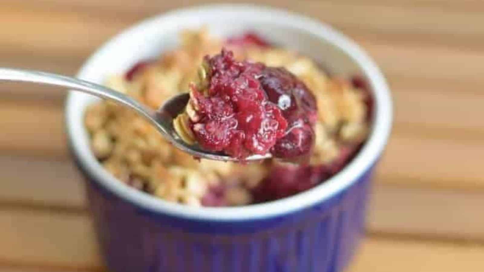 Image shows A spoon lifting berry oatmeal from a ramekin, showcasing the juicy fruit topping and golden-brown crust beneath.