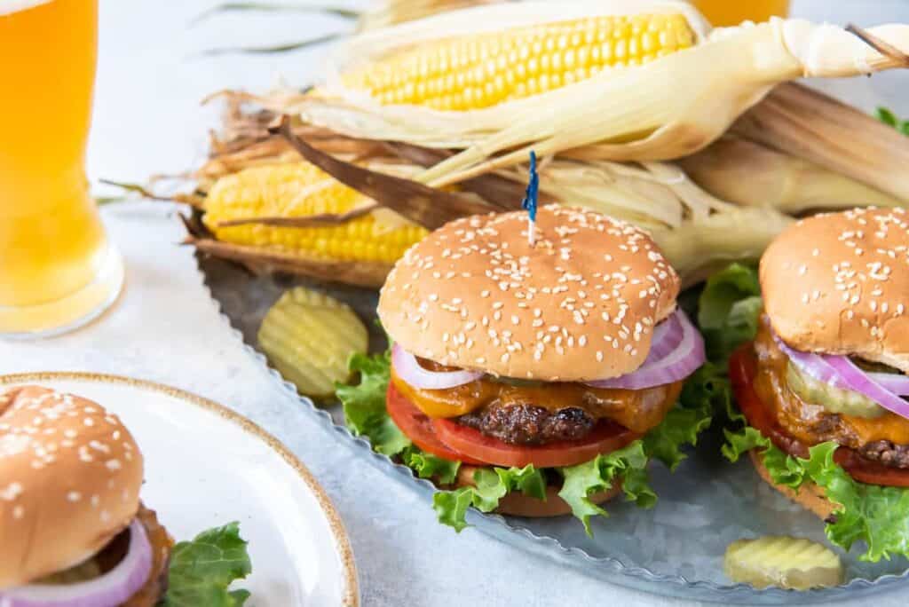 A plate with cheeseburgers garnished with lettuce, tomato, and onion, accompanied by corn on the cob and pickle slices. There are glasses of beer in the background.