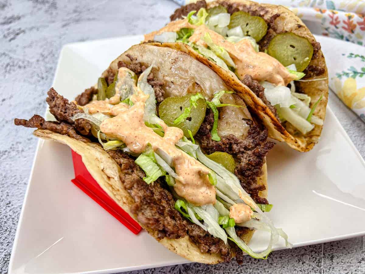 Two Big Mac tacos filled with ground beef, shredded lettuce, and pickles, and drizzled with a creamy sauce, placed on a white plate.
