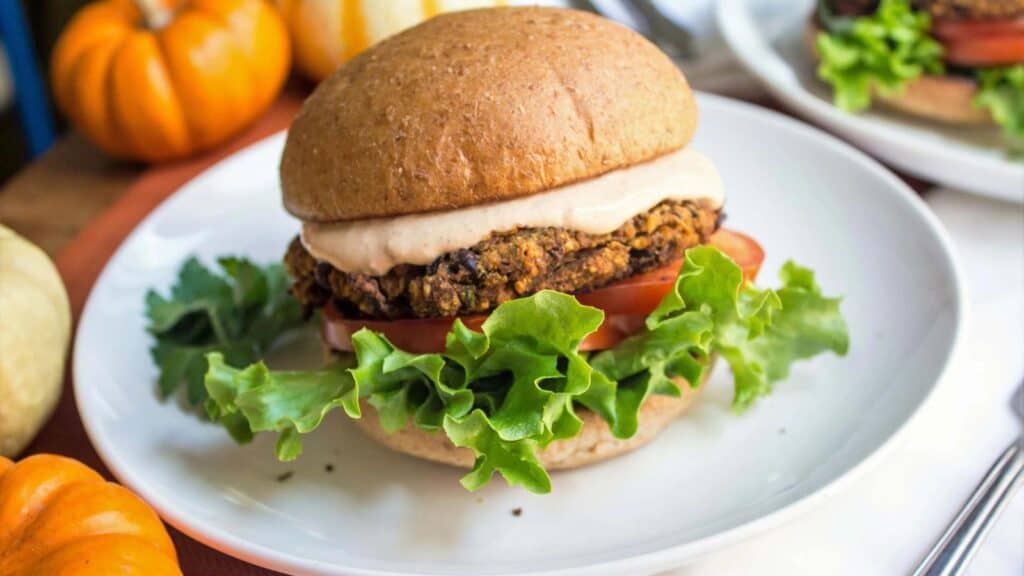 A veggie burger with lettuce, tomato, and sauce on a whole wheat bun, served on a white plate with small pumpkins in the background.