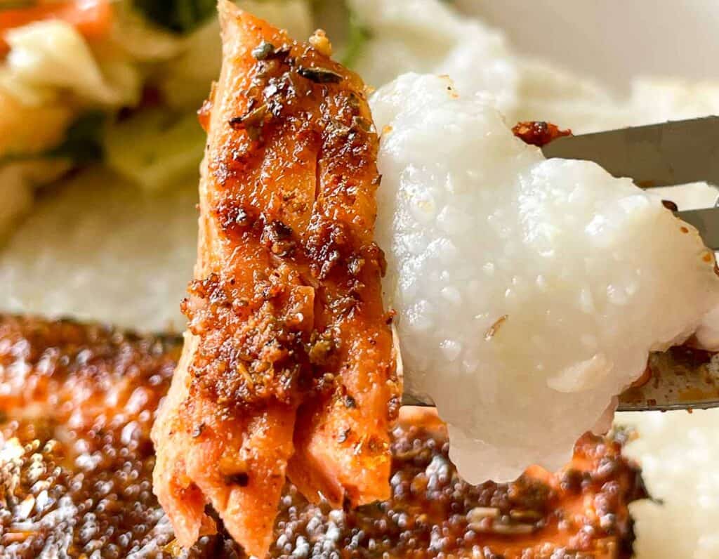 A close-up of a fork holding a piece of seasoned salmon and a portion of white grits, with some vegetables visible in the background.